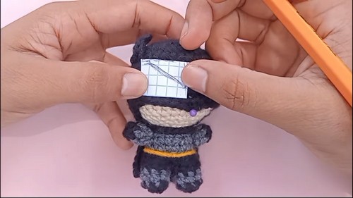 Hands pressing a small paper template onto the black cowl of a Batman amigurumi to mark placement for the felt eye pieces