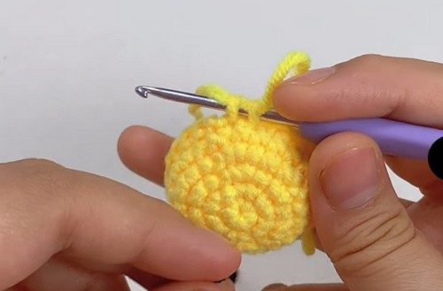 Hands using a purple crochet hook to add a keychain loop to a small round yellow crocheted bee body.