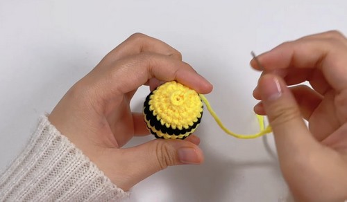 Hands holding a yellow and black crocheted bee body from the top, showing the stripe pattern in progress with a yarn tail extending to the right.