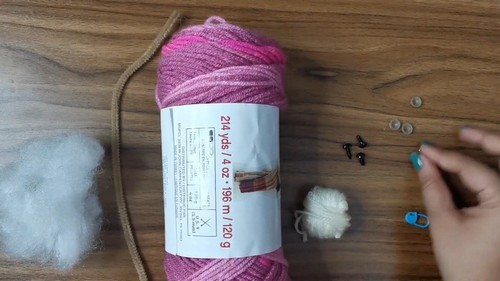 Supplies for the crochet cat heart pattern laid out on a wood surface including a skein of pink variegated yarn safety eyes fiberfill and a small crochet hook