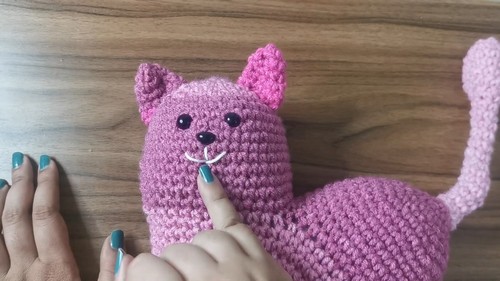Hands embroidering a white nose and mouth detail onto a finished pink crochet cat heart amigurumi face on a wooden surface
