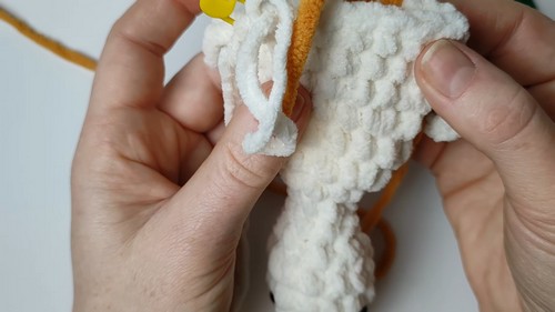 Hands holding a white chenille goose amigurumi mid-assembly, showing the long neck piece being joined to the body with a yellow crochet hook visible at the top.