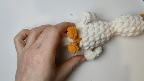Hand positioning three small orange crocheted foot pieces at the base of a white chenille goose amigurumi body before attachment.