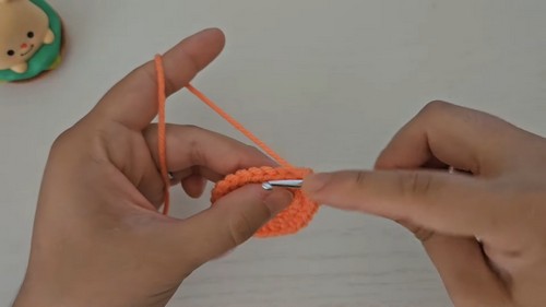 Hands working the foundation round of an orange hermit crab body piece using a metal crochet hook.