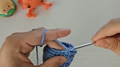 Hands crocheting the blue bucket shell piece using a metal hook, with the orange crab body visible in the background.
