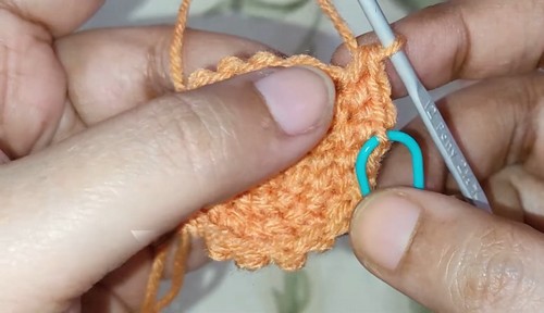 Hands using a white crochet hook and blue stitch marker to work the early rounds of a small orange crocheted deer snout piece.