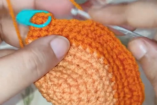 Hands holding a large stuffed orange crocheted deer head with a blue stitch marker and a second crochet hook visible at the side, mid-assembly.
