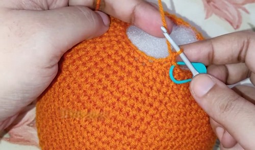 Hands using a blue crochet hook to work orange yarn stitches across the top of a stuffed deer head, with polyfill visible in the opening.