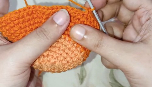 Hands joining a lighter orange crocheted snout piece to the main orange deer head body using a crochet hook, showing the color change seam.