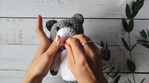 Hands attaching the white muzzle piece onto the front of a gray crocheted bear head with fingers pressing it into position