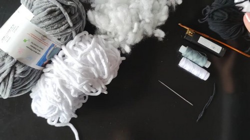 Supplies for the crochet bear pattern laid out on a dark surface including gray and white chenille yarn fiberfill stuffing crochet hooks a tapestry needle and black embroidery thread