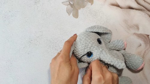 Hands positioning the face of a gray crochet elephant amigurumi showing blue safety eyes and a trunk during assembly