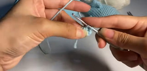 Hands using a tapestry needle to sew a fin piece onto the back of a light blue crochet tiger shark amigurumi in progress.