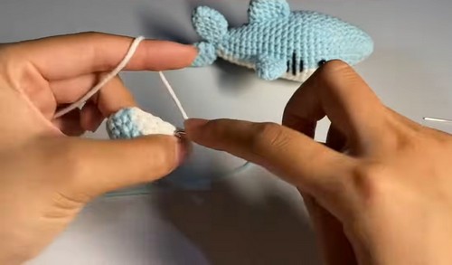 Hands attaching a small crocheted pectoral fin to the side of a light blue tiger shark amigurumi body using a tapestry needle.