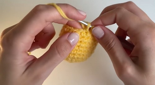 Hands working the upper rounds of a yellow crochet egg shape with a small hook.