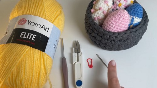Flat lay of a yellow YarnArt Elite yarn skein, crochet hook, scissors, and stitch marker beside a gray basket holding several decorated multicolored crochet Easter eggs.
