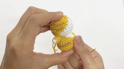 Hands holding a small yellow and white crocheted cat body and head joined together with a pin holding pieces in place during assembly