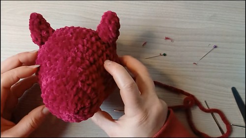 Hands pinning small crocheted ears onto a dark red dragon head with sewing pins placed around to position them before attaching