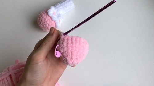 Hand holding a crocheted pink cow body piece with a crochet hook inserted, second piece visible in background