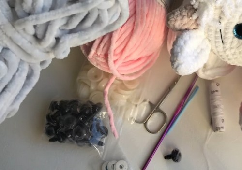Flat lay of supplies for crochet cow amigurumi — pink and white chenille yarn, safety eyes, scissors, crochet hooks, and a partially made doll