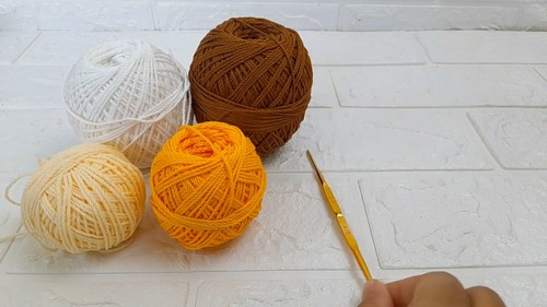 Flat lay of four yarn balls in white, cream, orange, and brown beside a gold crochet hook on a white brick background.