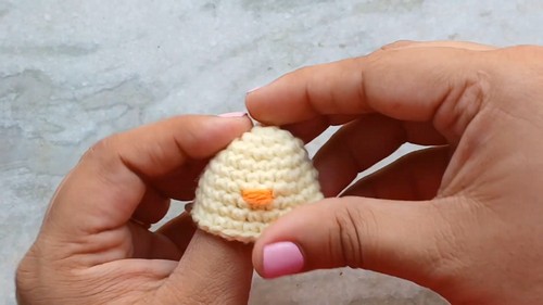 Hands holding a small dome shaped yellow crocheted chick piece showing an embroidered orange beak on the front before eyes are added