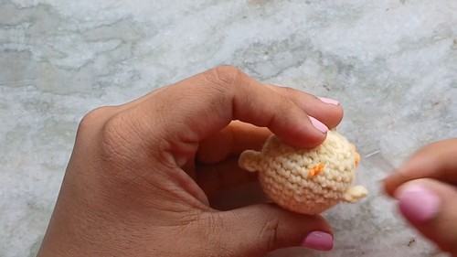 Hand holding a small crocheted yellow chick body with a tiny orange beak already attached showing the stage just before adding the eyes