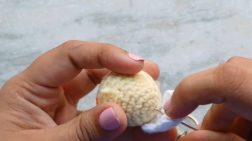 Hands using a tapestry needle to attach a white crocheted eggshell cap piece onto the top of a small yellow chick head