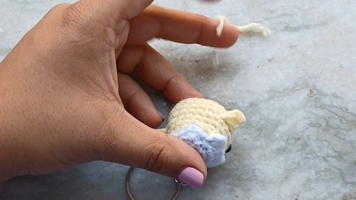 Hand holding a small crocheted yellow chick body sitting inside a blue egg base with a white eggshell top and a silver keyring attached at the bottom