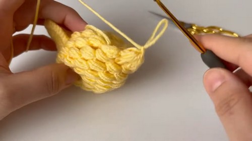 Hands shaping a yellow crocheted chicken body piece with a gold hook showing the rounded body form taking shape with multiple yarn tails visible