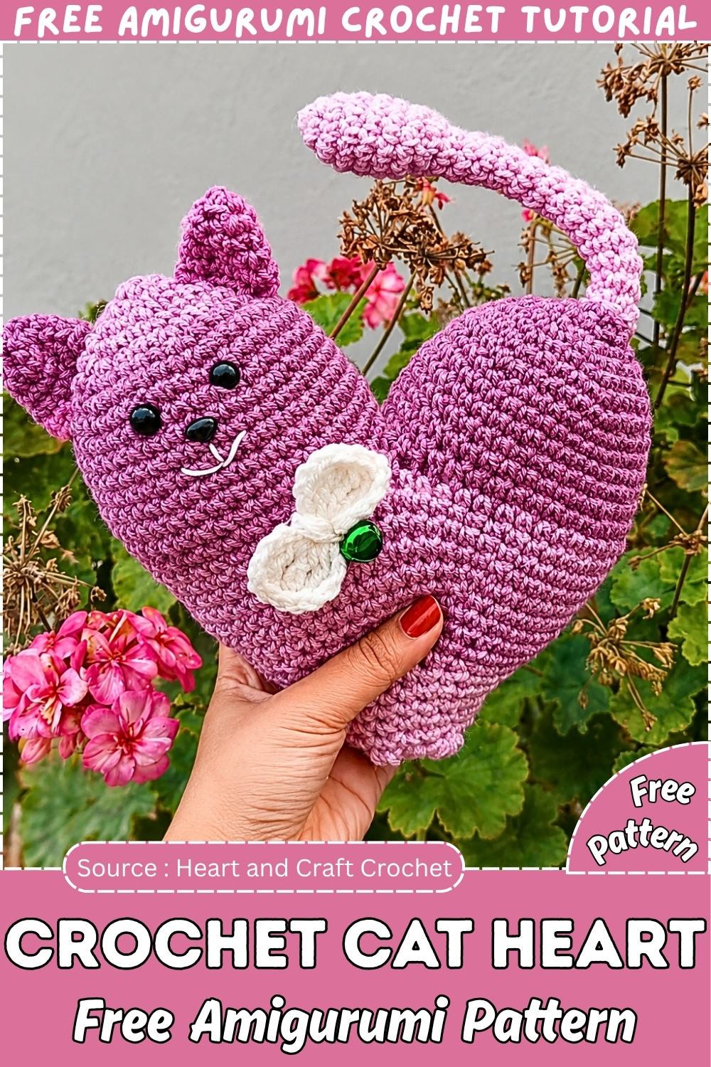 Finished purple crochet cat heart amigurumi with ears tail white bow and green gem detail held up against a garden background with pink geraniums