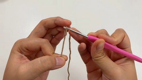 Hands using a pink crochet hook to form the first chain stitches in tan yarn at the very start of a capybara amigurumi piece.