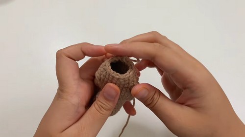 Hands holding a tan crocheted capybara body piece from above, showing the open top before stuffing and closing.