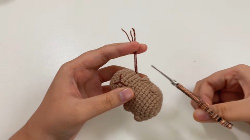 Hands holding a small tan crocheted capybara head with dark brown ear yarn tails and a decorative crochet hook resting nearby on a white surface.