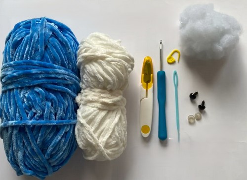 Supplies laid out for a crochet mini shark pattern including blue and white chenille yarn, crochet hooks, stitch markers, a tapestry needle, polyester stuffing, and black safety eyes.