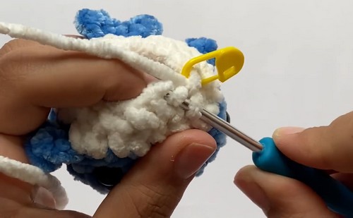 Hands working the color join between blue and white chenille yarn on a crochet mini shark amigurumi body using a blue hook and yellow stitch marker.