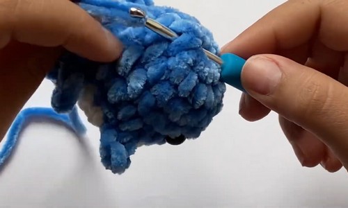 Hands attaching a blue chenille fin to the top of a small crochet mini shark body using a tapestry needle.