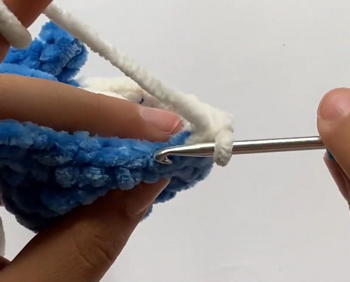 Hands crocheting the white chenille belly section onto the base of a blue mini shark amigurumi body using a metal hook.