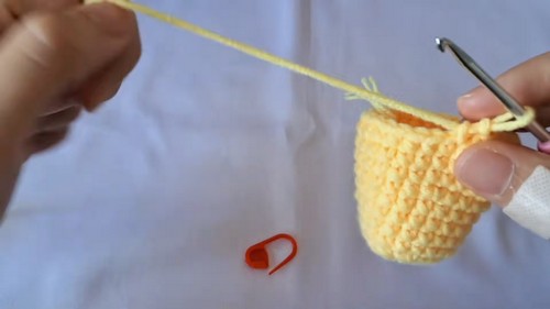 A partly worked yellow crochet baby chick body on a hook with a red stitch marker beside it.