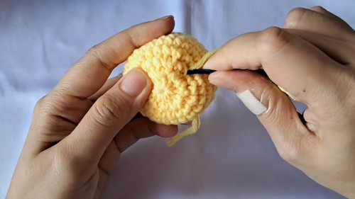 A safety eye being inserted into a stuffed round yellow crochet baby chick head.