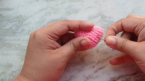 Hands using a needle to close and finish a small crocheted pink heart amigurumi showing the seaming step