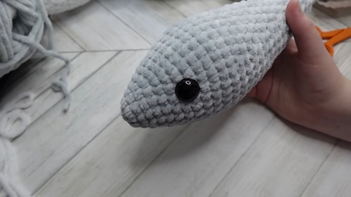 Hand holding the stuffed gray chenille head of a crochet shark amigurumi showing a large black safety eye positioned on the side.