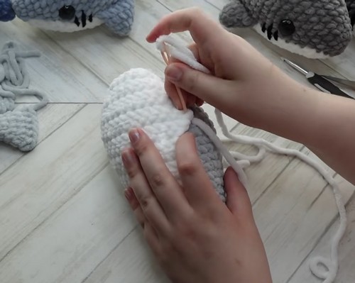 Hands sewing the white chenille belly panel onto the gray body of a crochet shark amigurumi using a tapestry needle.