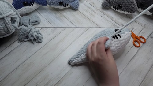 Hand smoothing the flat belly piece of a chenille crochet shark amigurumi on a wooden floor with additional finished sharks visible in the background.
