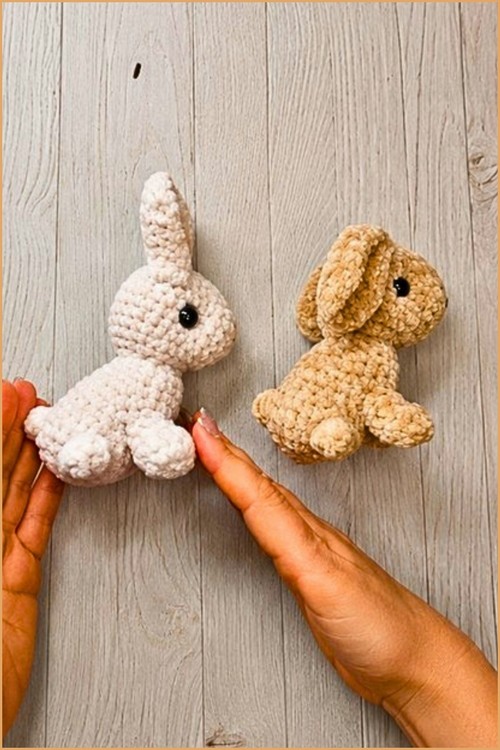 A white velvet crochet bunny with upright ears and a tan floppy-eared velvet bunny placed side by side on a gray wood surface, with two hands shown touching the white bunny from below.