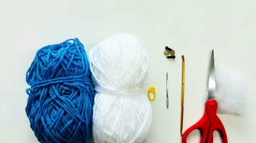 Flat lay of supplies for a crochet whale shark pattern including blue and white yarn, safety eyes, a crochet hook, scissors, a tapestry needle, and stuffing.