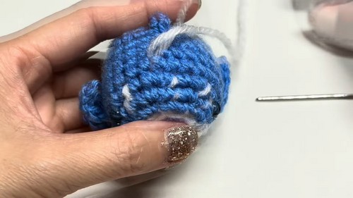 Hands holding a partially completed blue and white crochet whale shark body during assembly, with a tapestry needle visible in the background.
