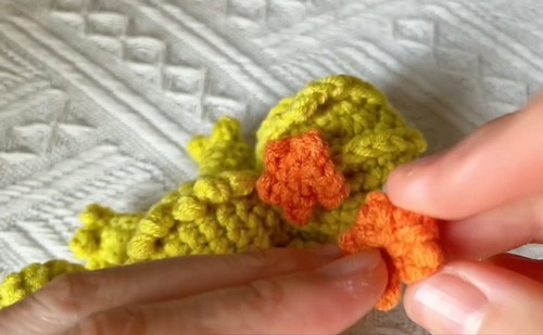 An orange crocheted crest being held against the back of a yellow crochet chameleon body.