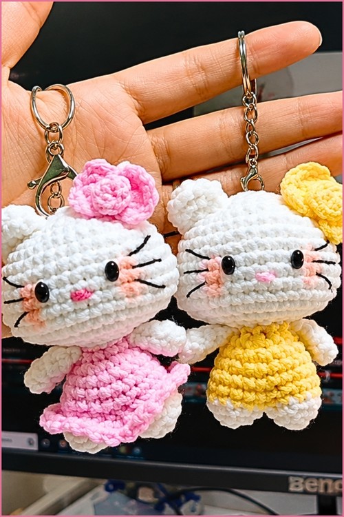 Two finished mini kitten amigurumi keychains, one in pink and one in yellow, held in an open palm with silver keyring hardware attached.
