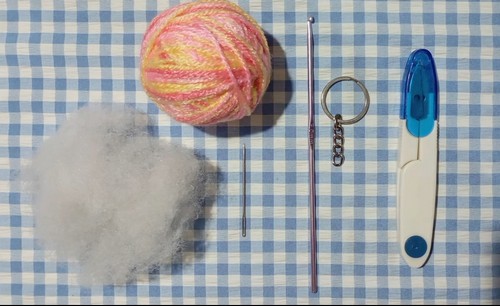 Materials laid out on a blue gingham surface including a ball of pink and yellow variegated yarn, polyfill stuffing, a crochet hook, tapestry needle, keychain ring, and stitch marker.